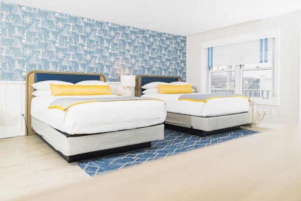 A bright room with two beds, yellow accents, nautical wallpaper, blue rug, white walls, and large window.