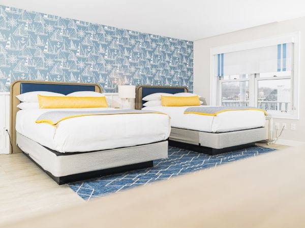 A bright room with two beds, yellow accents, nautical wallpaper, blue rug, white walls, and large window.