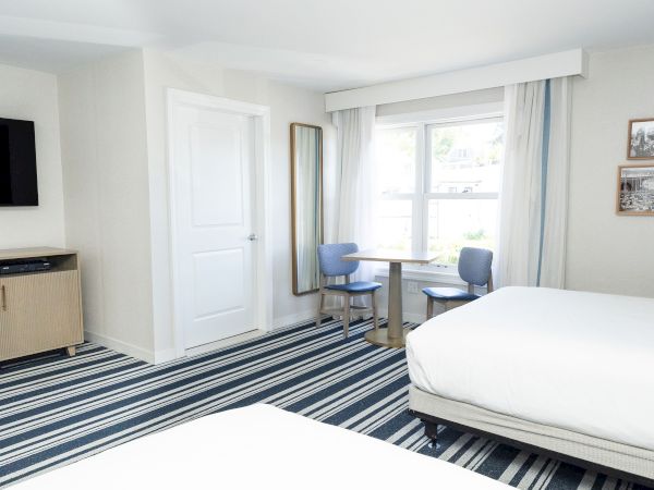 Bright hotel room with a small table, two blue chairs, curtains and window, plus a TV, cabinet, and framed photos.
