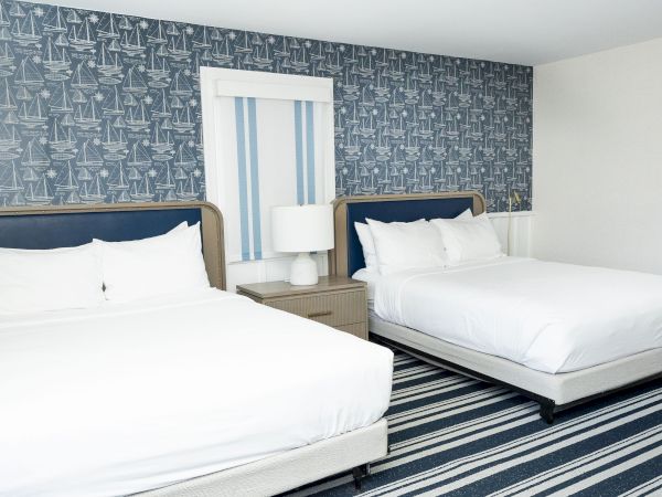 A nautical-themed guest bedroom with two blue headboards, white bedding, and sailboat wallpaper behind the beds.