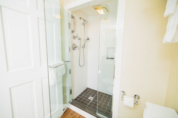 The image shows a modern bathroom with a glass-enclosed shower, multiple showerheads, a gray tiled floor, a towel rack, and a toilet paper holder.