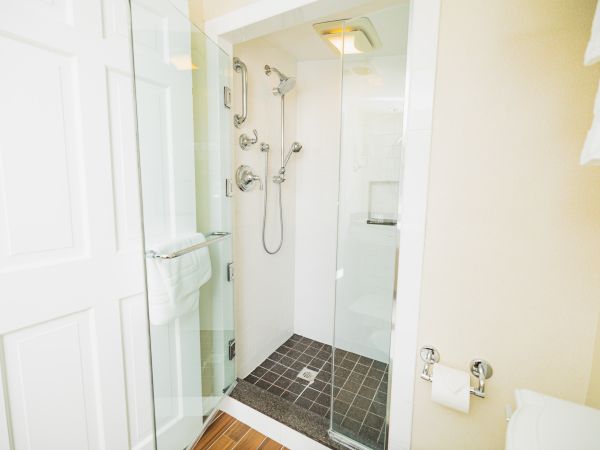 The image shows a modern bathroom with a glass-enclosed shower, multiple showerheads, a gray tiled floor, a towel rack, and a toilet paper holder.