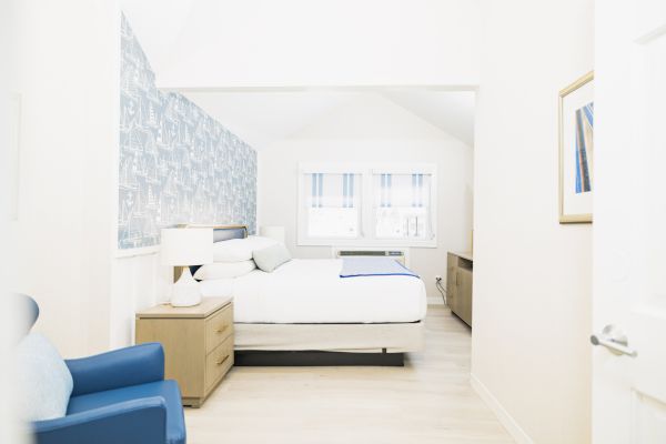 A bright, minimalist bedroom with a bed, blue armchair, wooden nightstands, lamps, and a window with striped curtains. The wall has patterned wallpaper.