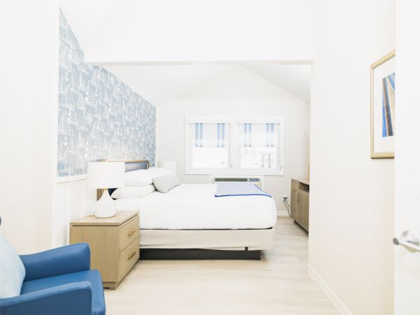A bright, minimalist bedroom with a bed, blue armchair, wooden nightstands, lamps, and a window with striped curtains. The wall has patterned wallpaper.