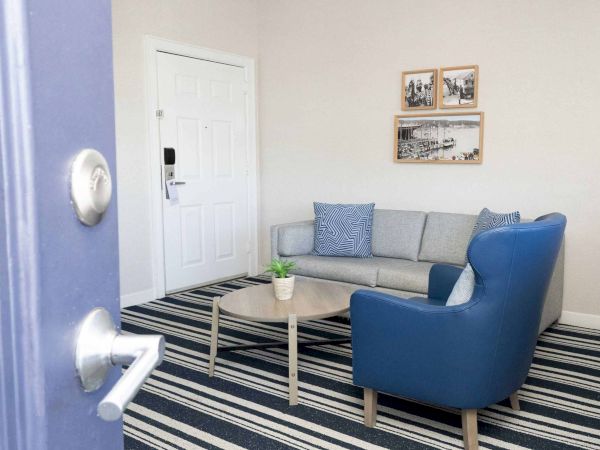 A bright living room corner with a white door, gray sofa, blue armchair, striped rug, framed photos, and a small plant.