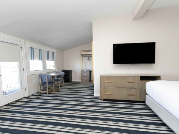 A bright hotel room with a bed, wall-mounted TV, striped window blinds, a small dining table, and a patterned carpet.