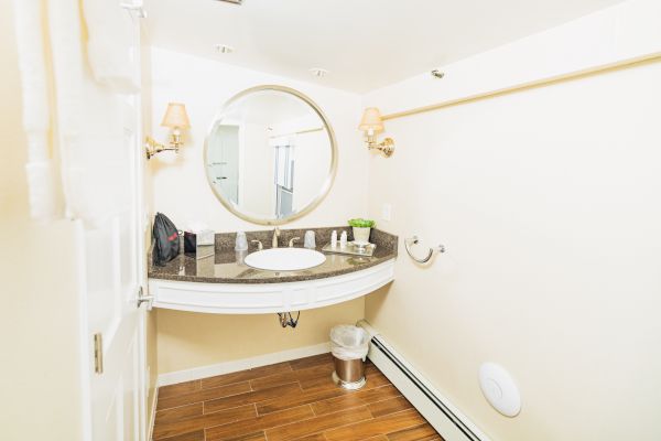 A modern bathroom with a round mirror above a countertop sink, wall lamps, towel rack, wooden flooring, and minimal decor.