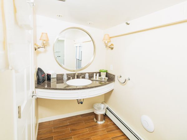 A modern bathroom with a round mirror above a countertop sink, wall lamps, towel rack, wooden flooring, and minimal decor.