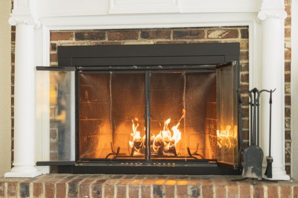 This image shows a lit fireplace with a metal screen and glass doors, flanked by white columns. Fireplace tools are placed on the right side.