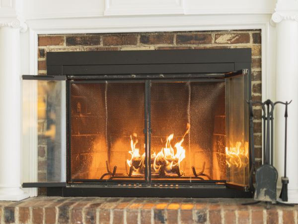 This image shows a lit fireplace with a metal screen and glass doors, flanked by white columns. Fireplace tools are placed on the right side.