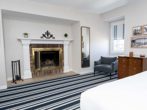 A bright living room with a brick fireplace, striped rug, blue chairs, a dresser, and framed photos on the wall.