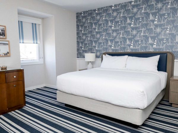 A bright bedroom with a large bed, white bedding, wooden dressers, striped rug, and navy nautical wallpaper covering the walls.