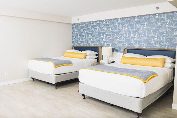 The image shows a hotel room with two neatly made beds, yellow and gray accents, and a blue nautical-themed wallpaper on the wall.