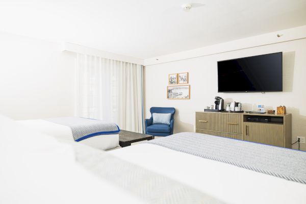 A hotel room with two beds, a wall-mounted TV, a blue chair, and a cabinet with a coffee maker and amenities.