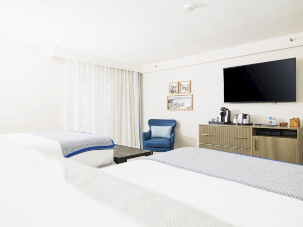 A hotel room with two beds, a wall-mounted TV, a blue chair, and a cabinet with a coffee maker and amenities.