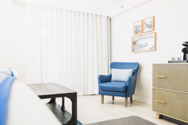 A bright room with a blue armchair, light-colored curtains, framed pictures, a wooden table, and a dresser.