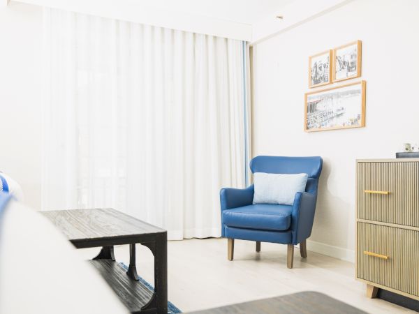 A bright room with a blue armchair, light-colored curtains, framed pictures, a wooden table, and a dresser.