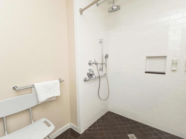 A bright bathroom showing a wall-mounted shower with handheld sprayer and hose above a tiled bath area, with toiletries on the wall.