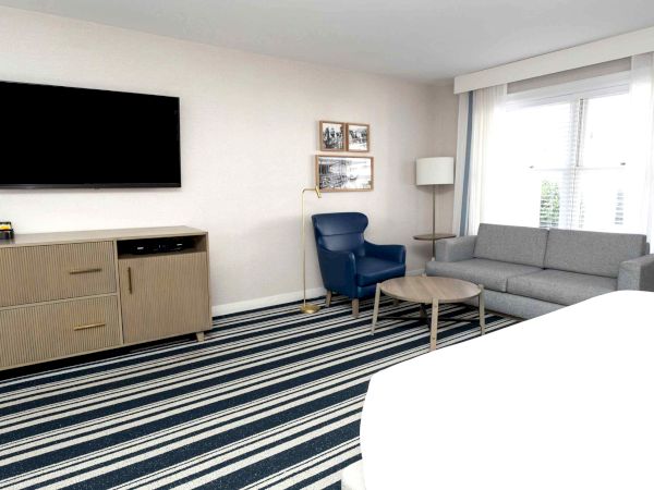 A modern living room with a wall TV, wood media cabinet, gray sofa and blue armchair, floor lamp, and large bright window with blinds.