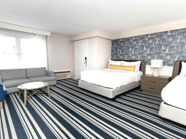 A bright hotel room with two twin beds, gray sofa, striped rug, and nautical boat-and-anchor wallpaper.