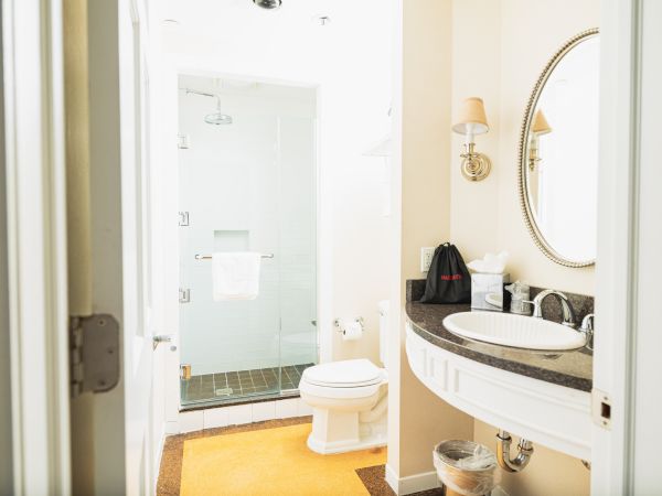 The image shows a bathroom with a glass-enclosed shower, a toilet, a sink with a mirror, a black travel bag, and a towel hanging on a bar.