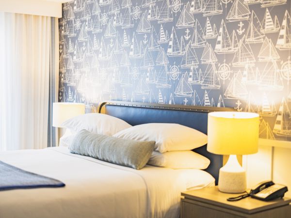 A neatly made bed with a blue headboard, two bedside lamps, nautical-themed wallpaper, and a bedside table with a phone and notepad.