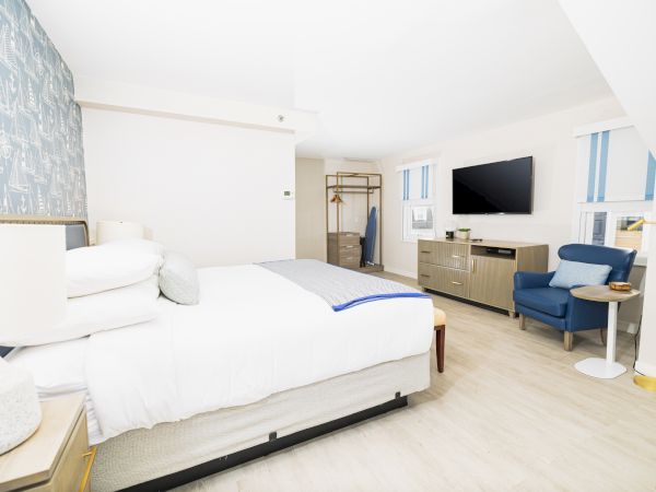The image shows a bright, modern hotel room with a bed, flat-screen TV, dresser, chair, side table, and hanging space for clothes.