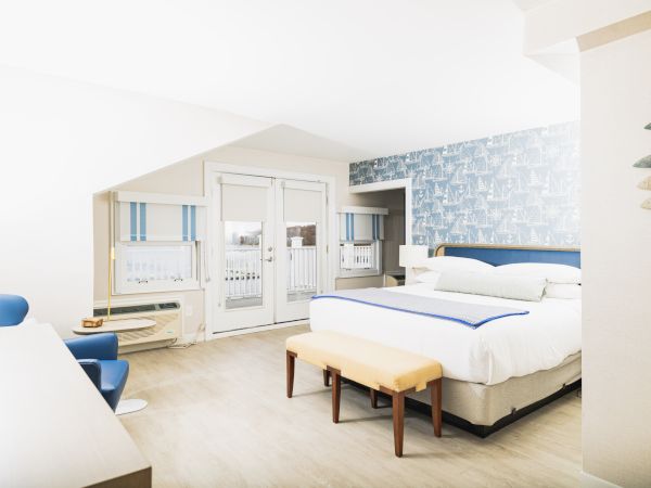 A well-lit hotel room with a bed, a bench, blue chairs, a desk, windows with shades, patterned wall, and decor featuring hanging fish artwork ends the sentence.