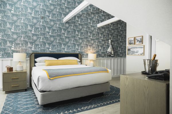 A cozy bedroom with a blue patterned accent wall, bed with yellow and gray bedding, two bedside tables with lamps, framed artwork, and a small dresser.