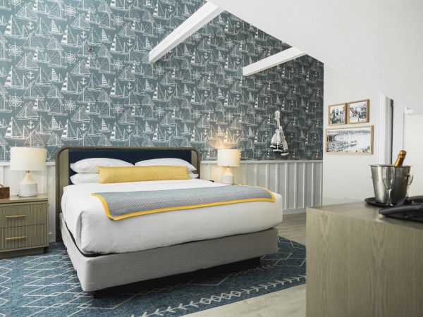 A cozy bedroom with a blue patterned accent wall, bed with yellow and gray bedding, two bedside tables with lamps, framed artwork, and a small dresser.