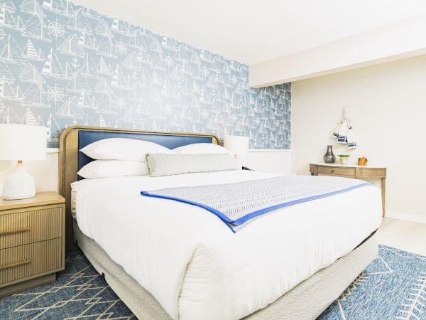 A stylish bedroom with a nautical theme features a large bed, blue wallpaper with sailboats, a nightstand, and a dresser with decorations.