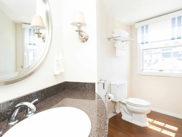 A bright bathroom with a granite vanity, chrome light fixtures, striped window shades, and a white toilet.