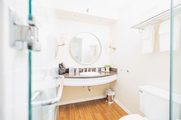 A modern bathroom with a round mirror, sink, countertop, toiletries, towels, a trash can, and a toilet view on wooden flooring.