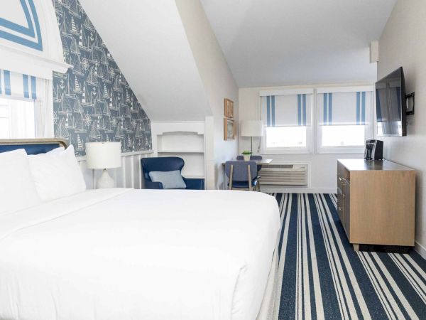 A modern hotel room with a white bed, chair and desk, striped carpet, and sailboat-themed wall behind the pillows.