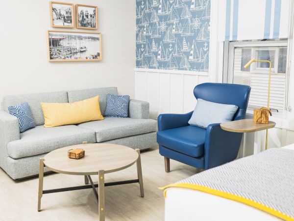A cozy living room with a blue armchair, gray sofa, wooden coffee table, and nautical-themed decor in blue and yellow tones, plus wall art.