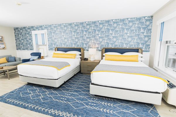 The image shows a bright bedroom with two double beds, nautical-themed decor, a blue accent wall, a patterned rug, and some seating.