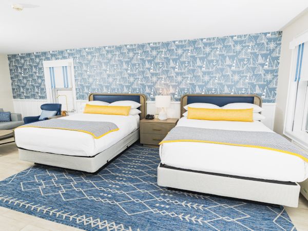 The image shows a bright bedroom with two double beds, nautical-themed decor, a blue accent wall, a patterned rug, and some seating.