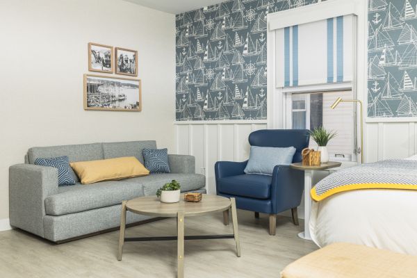 A cozy living room with a gray sofa, blue armchair, wooden coffee table, and nautical-themed wallpaper, featuring wall art and modern decor.