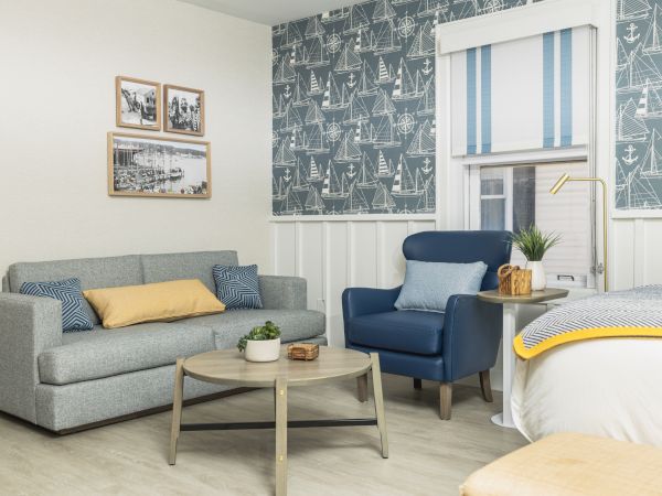 A cozy living room with a gray sofa, blue armchair, wooden coffee table, and nautical-themed wallpaper, featuring wall art and modern decor.
