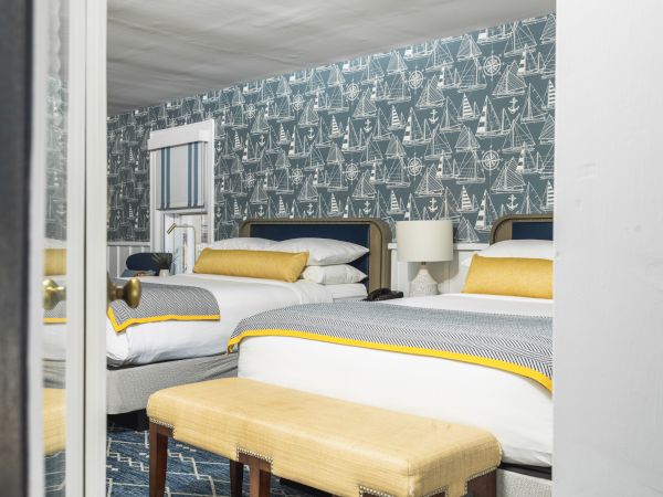 A cozy bedroom with two neatly made beds, yellow accents, a nautical-themed wallpaper, a small bench, a side table, and a lamp is shown.