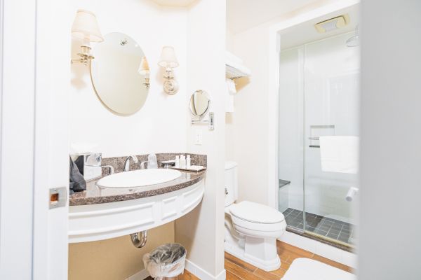 The image shows a clean and well-lit bathroom with a circular mirror, sink, toilet, and a glass-enclosed shower area, featuring modern amenities.