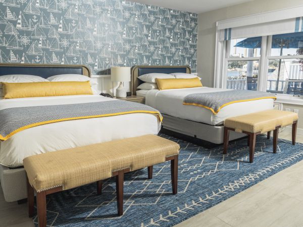A bedroom with two double beds, yellow and grey bedding, bench seats, patterned wallpaper, a blue rug, a nightstand, and a window.