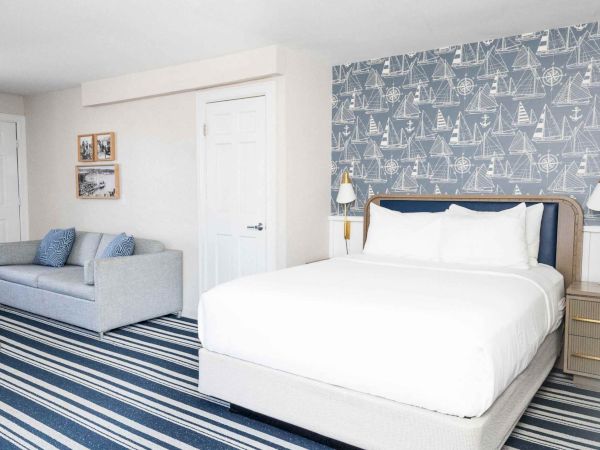 A bright bedroom with a large bed, cozy couch, nautical boat-pattern wallpaper, striped rug, and bedside lamps.