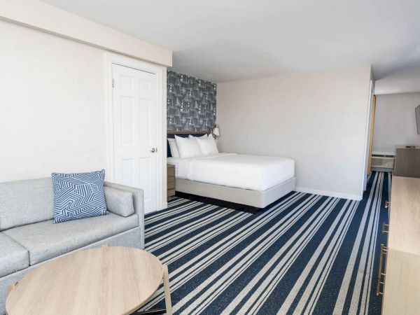 A modern hotel room with a bed, gray sofa, striped carpet, patterned wall art, and a TV console.