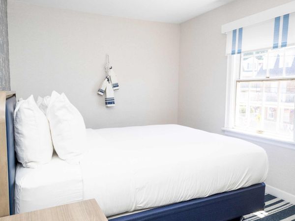 A bright bedroom with a neatly made white bed, blue-and-white striped curtains at a window, and simple wall decor.