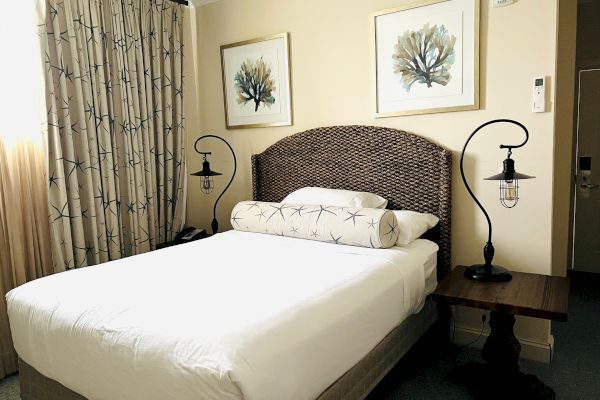 A neatly made bed with a woven headboard, flanked by two nightstands with industrial-style lamps, and two framed artworks above.