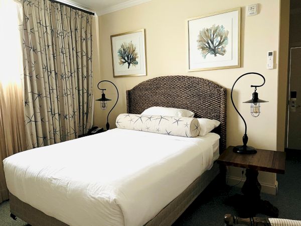 A neatly made bed with a woven headboard, flanked by two nightstands with industrial-style lamps, and two framed artworks above.