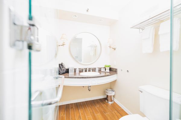 The image shows a bathroom with a round mirror, sink, towels, a toilet, a trash bin, and toiletries on the counter, with wooden flooring.