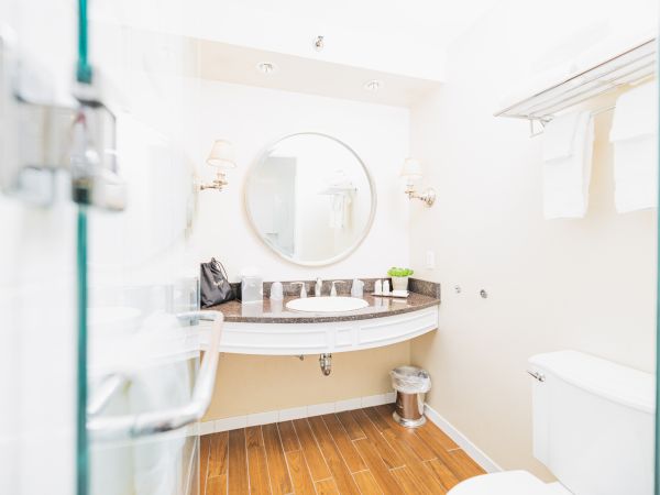 The image shows a bathroom with a round mirror, sink, towels, a toilet, a trash bin, and toiletries on the counter, with wooden flooring.