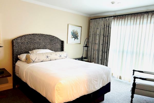 A hotel room with a bed, side table, large window with curtains, decorative painting, and an armchair.